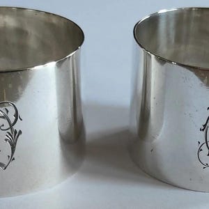 May include: Two silver napkin rings with a polished finish. Each ring features an engraved monogram with ornate floral detailing. The rings are cylindrical with a slightly angled top edge, reflecting light.