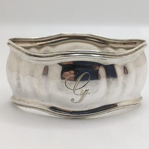 May include: A silver-colored, wavy-edged bowl with a curved body. The bowl has a reflective surface and a decorative letter "G" engraved on the front. The bowl is likely made of metal and is designed for decorative or functional use.