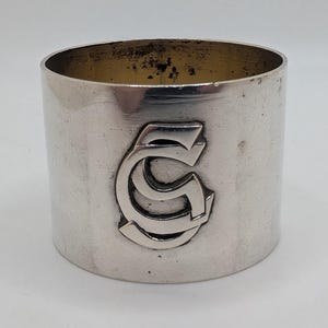 May include: A silver napkin ring, cylindrical in shape, featuring a raised "SC" monogram. The interior shows slight tarnishing. The metal has a polished finish. The monogram is centrally placed on the front of the ring.