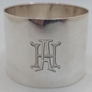 Può includere: Un portatovagliolo d'argento con finitura lucida. L'anello presenta un monogramma inciso con le lettere "AH" in un carattere decorativo. L'anello cilindrico è realizzato in metallo lucido e riflettente.