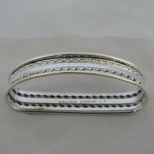 Puede incluir: Un servilletero plateado de forma rectangular. Presenta un diseño trenzado en los bordes superior e inferior, con una banda blanca lisa en el centro. El texto "WALLACE STERLING" es visible.