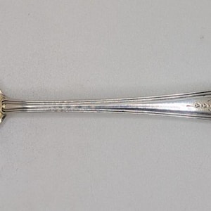 May include: A silver-toned metal spoon featuring a round bowl and a slender handle. The handle is decorated with an embossed floral pattern and a central monogram detail. The spoon has a polished finish with visible light reflections on the bowl.