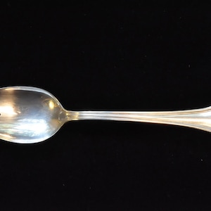 Op de afbeelding: Een zilveren serveerlepel met een gleuf, drietandig uiteinde. De lepel heeft een lange, slanke steel met een decoratief ontwerp aan het uiteinde. De lepel staat tegen een zwarte achtergrond.