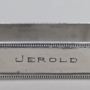 May include: A rectangular silver napkin ring with the name "JEROLD" engraved on the front. The edges are detailed with a beaded design. The ring is designed to hold a folded napkin.