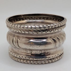 Può includere: Un anello portatovagliolo d'argento con un design dettagliato. L'anello ha un bordo a forma di corda nella parte superiore e inferiore, con una sezione centrale strutturata e curva. L'anello ha iniziali e segni incisi. Il design generale è ornato e classico.