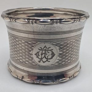 May include: A silver napkin ring with an engraved monogram. The ring features a wavy pattern and horizontal lines. The top and bottom edges have a decorative beaded design. The monogram is in a circular frame.
