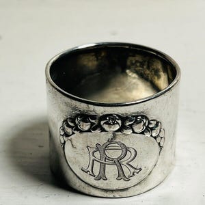 May include: A silver napkin ring with an engraved design. The ring features a central oval with the intertwined letters "R" and "R" and is surrounded by a floral motif. The ring has a cylindrical shape.