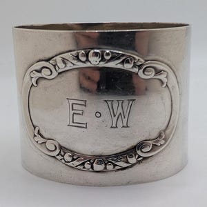 May include: A silver napkin ring with an oval cartouche bearing the engraved initials "E.W." The cartouche is framed by an ornate, scrolling design. The ring is cylindrical and has a polished finish.