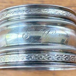 May include: A silver napkin ring with an engraved monogram. The ring features decorative scrollwork and a rope-like texture. The ring is made of a shiny, reflective metal.
