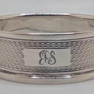 May include: A silver napkin ring with a textured wave pattern around the centre. The ring has a rectangular cartouche with the engraved initials "BS". The top and bottom edges are smooth and rounded.