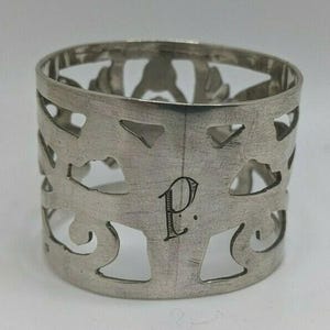 Pode incluir: Um porta-guardanapo prateado com um design vazado com formas florais e geométricas estilizadas. A letra "P" está gravada na frente. O anel é feito de metal brilhante.