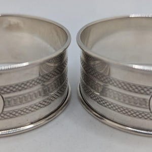 Puede incluir: Un par de servilleteros circulares de plata con un acabado metálico pulido. Cada anillo está decorado con una banda horizontal texturizada y un medallón circular central grabado con la letra cursiva G en la parte frontal.