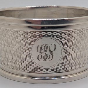 May include: A silver napkin ring with a textured wave pattern around the center. The ring has a circular monogram in the center and a smooth rim at the top and bottom. The ring is approximately 2 inches in diameter.