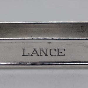 Puede incluir: Un servilletero rectangular plateado con bordes redondeados. La palabra "LANCE" está grabada en el centro del anillo. El anillo tiene una superficie pulida y reflectante.