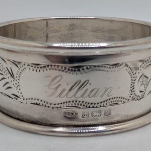May include: A silver napkin ring, engraved with the name "Gillian" in cursive script. The ring features decorative floral and leaf patterns. Hallmarks are visible on the base of the ring.