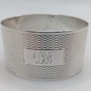 May include: A cylindrical silver napkin ring featuring a fine, repeating engine-turned cross-hatch pattern across its exterior surface. A smooth, rectangular central cartouche is engraved with the stylized cursive initials DB in the center.