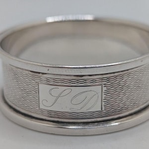 Könnte beinhalten: Ein silberner Serviettenring mit einem strukturierten Wellenmuster und einer rechteckigen Kartusche mit den Initialen "S D". Der Ring ist kreisförmig und hat eine polierte Oberfläche.
