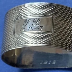 May include: A silver napkin ring with a textured, diamond-patterned surface. The ring features an engraved rectangular plaque with the initials "F B" and the date "12.9.43". The year "1918" is also engraved on the ring.