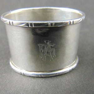 Può includere: Un portatovagliolo d'argento con monogramma. L'anello è cilindrico con linee decorative incise attorno ai bordi superiore e inferiore. Il monogramma è inciso sulla parte anteriore.