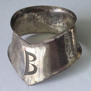 Pode incluir: Um colar de metal prateado com uma textura martelada. O colar tem uma faixa larga e curva que se estreita em uma ponta na frente. Uma grande letra "B" é recortada do metal na frente.
