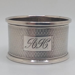 Può includere: Un anello portatovagliolo d'argento con un motivo a onde testurizzate e un cartiglio rettangolare. Il cartiglio presenta le iniziali incise "A.H.C.". L'anello ha una finitura lucida e un design classico.