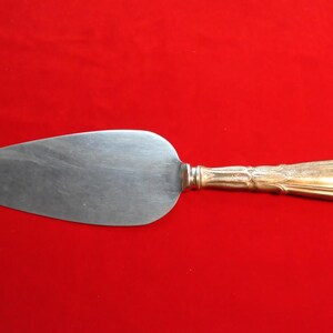 May include: A silver cake server with a gold-colored handle. The server has a wide, flat blade and a decorative handle. The handle has an ornate design. The server is set against a red background.