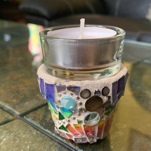 May include: A small, white candle in a glass holder sits on top of a colorful mosaic candle holder. The mosaic is made of small, colorful tiles and has a rainbow pattern.
