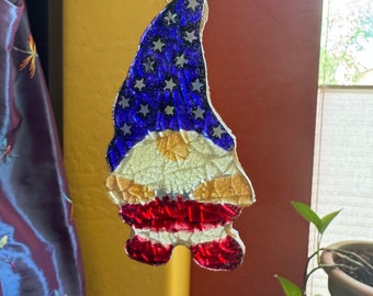 May include: A patriotic gnome suncatcher with a blue starry hat, a gold body, and red striped legs. The suncatcher is hanging from a beaded strand. The gnome is made of stained glass and is hanging in front of a window.