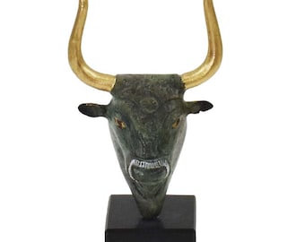 Pode incluir: Uma escultura de cabeça de touro de bronze com chifres dourados sobre uma base preta. Os olhos do touro são verdes e a boca está aberta.