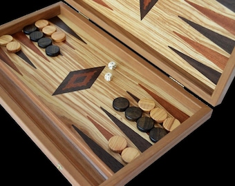 May include: A wooden backgammon set with a hinged lid, open to reveal the game board. The board is made of light and dark wood with a diamond pattern in the center. There are white and black game pieces on the board, as well as two white dice.