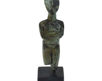 May include: A small, green bronze statue of a standing figure with arms crossed. The figure is on a black base.