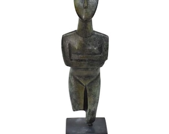May include: A bronze sculpture of a standing figure with arms crossed. The figure has a simplified, stylized form with a rounded head and a long, slender body. The sculpture is mounted on a black base.