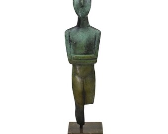 May include: A bronze sculpture of a standing figure with arms crossed. The figure has a simplified, stylized form and is mounted on a dark brown marble base.
