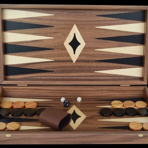 May include: A wooden backgammon board set featuring a walnut finish with inlaid black and light wood triangles. The board includes a set of round wooden checkers in light and dark tones, two white dice, one black doubling cube, and a brown cup.