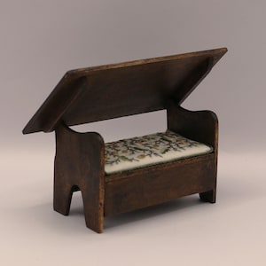 May include: A miniature wooden bench in dark brown, featuring a hinged lid. The seat cushion is patterned with green and yellow floral motifs on a white background. A small piece of furniture.