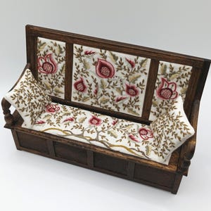May include: A miniature wooden bench with embroidered floral patterns. The dark brown frame supports a back panel and seat cushion decorated with red and white floral designs. Two matching pillows are included.