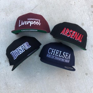 May include: Four baseball caps with team logos. A maroon cap with white text that says "Merseyside Liverpool", a black cap with red text that says "Arsenal", a black cap with white text that says "Hotspur", and a navy blue cap with white text that says "Chelsea Blues of Stamford Bridge".