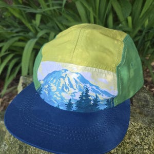 May include: A five-panel cap featuring a blue flat brim and a crown with lime green side panels. The front panel displays a graphic illustration of a snow-capped mountain peak above a forest of dark green pine trees against a light sky.