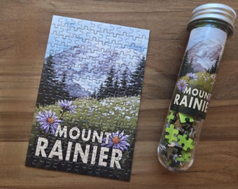 May include: A Mount Rainier puzzle with a mountain landscape scene, including trees, flowers, and a mountain range. The puzzle is next to a clear tube filled with puzzle pieces, also featuring the Mount Rainier design. The text "Mount Rainier" is visible.