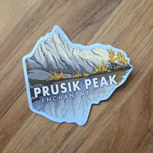 May include: A die-cut sticker featuring a mountain landscape with the text "PRUSIK PEAK ENCHANTMENTS." The design includes a mountain range, a reflective lake, and trees in shades of gray, yellow, and blue, with a light blue border.