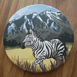 May include: Round coaster featuring a zebra walking through a grassy field with a mountain range in the background. The zebra has black and white stripes. The mountains are green and gray, and the sky is light blue.