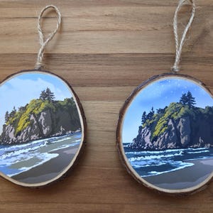 May include: Two round wooden ornaments with a coastal landscape design. Each ornament features a detailed illustration of a rocky island with trees, waves, and a beach. The ornaments have a natural wood edge and a jute twine loop for hanging.