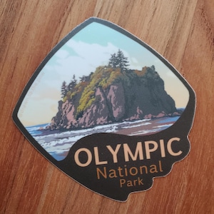 May include: A sticker featuring a vintage-style illustration of a rocky island with trees and a beach. The text "OLYMPIC National Park" is printed in brown and orange on a black background.
