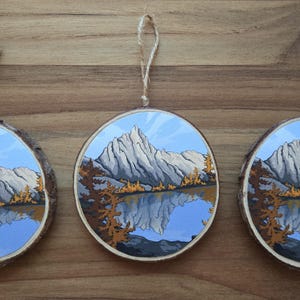 May include: Three round wooden ornaments with a painted mountain landscape scene. Each ornament features a gray mountain range reflected in a blue lake, with fall-colored trees. They have twine hangers.