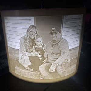 May include: A family portrait displayed in a light-up, curved-edge frame. The image shows a smiling woman, a baby in overalls, and a man in a baseball cap. The frame has a warm, inviting glow, perfect for a personalized gift.