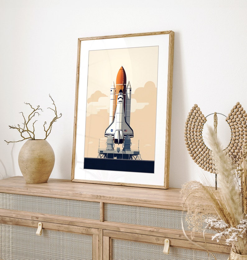 Pu&ograve; includere: Stampa artistica incorniciata con un'illustrazione stilizzata di uno space shuttle su una piattaforma di lancio. L'opera d'arte ha un'estetica retr&ograve; con una tavolozza di colori arancione, bianco e blu su uno sfondo beige. La stampa &egrave; in una cornice di legno.