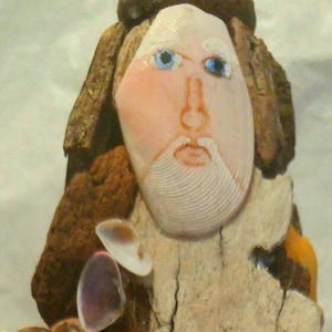 May include: A handcrafted sculpture of a bearded man's face, created from seashells and driftwood. The face is made from a large, light-colored shell, with blue eyes and a drawn-on beard. The hair and beard are made from brown driftwood.