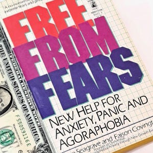 May include: A paperback book titled Free From Fears by Ann Seagrave and Faison Covington, featuring a grid-patterned cover with red and purple text, positioned next to a one-dollar bill showing the portrait of George Washington.