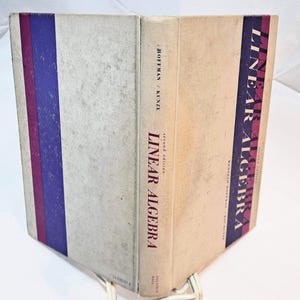 May include: A hardcover textbook titled Linear Algebra, second edition, by Hoffman and Kunze. The cover is off-white with a vertical purple and maroon stripe on the left side and a dark purple band on the right featuring the title in white text.