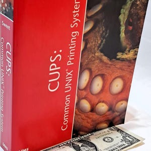 May include: A technical book titled CUPS: Common UNIX Printing System by Michael R. Sweet, featuring a red cover with an octopus tentacle graphic. A one-dollar bill is placed in front of the book to provide a visual scale for its size.